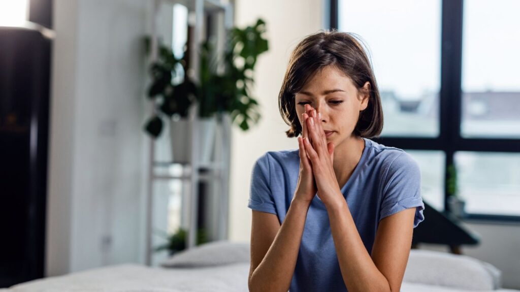 Woman sitting indoors with hands clasped, looking worried