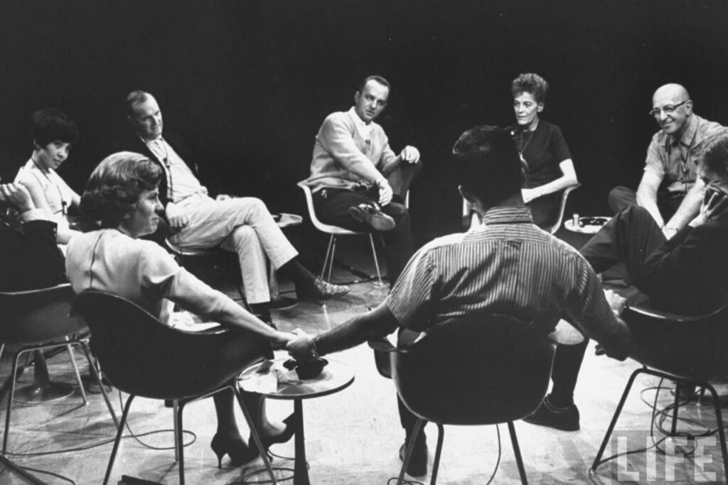 Black-and-white photo of a group of adults seated in a circle, holding hands during a group therapy or discussion session.