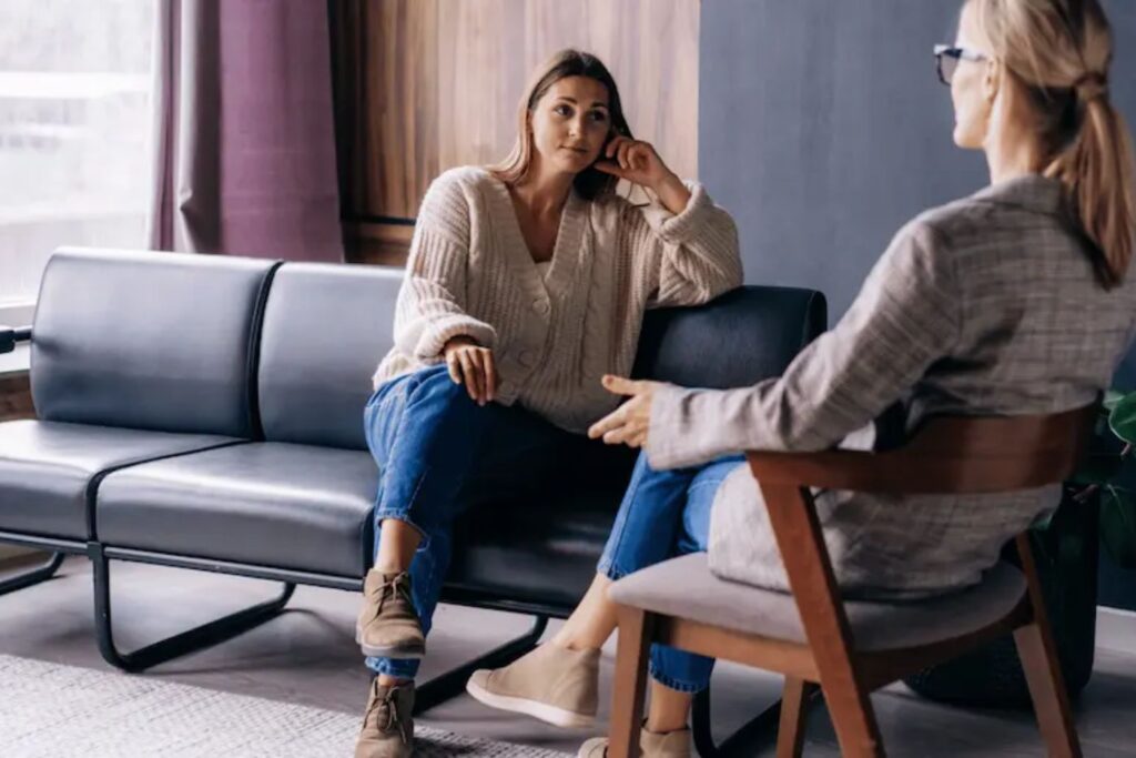 Two women seated across from each other in a counseling setting, engaged in conversation, one listening attentively while the other speaks.